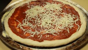 Sprinkling shredded mozzarella cheese over a red tomato sauce base on stretched pizza dough for a homemade pepperoni pizza