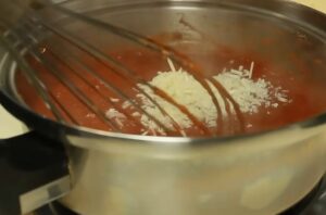 Whisking shredded parmesan cheese into simmering red tomato sauce in a stainless steel pot for homemade pepperoni pizza